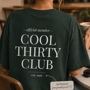 May include: A dark green short-sleeved t-shirt featuring white text on the back that reads - official member - COOL THIRTY CLUB and est. 1996. A small rectangular graphic with an arrow points to the text with the words Auf Wunsch personalisiert.
