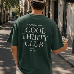 May include: A dark green short-sleeve t-shirt featuring white text on the back that reads - official member - COOL THIRTY CLUB est. 1996. The shirt is paired with a beige baseball cap and light-colored trousers, viewed from the back.
