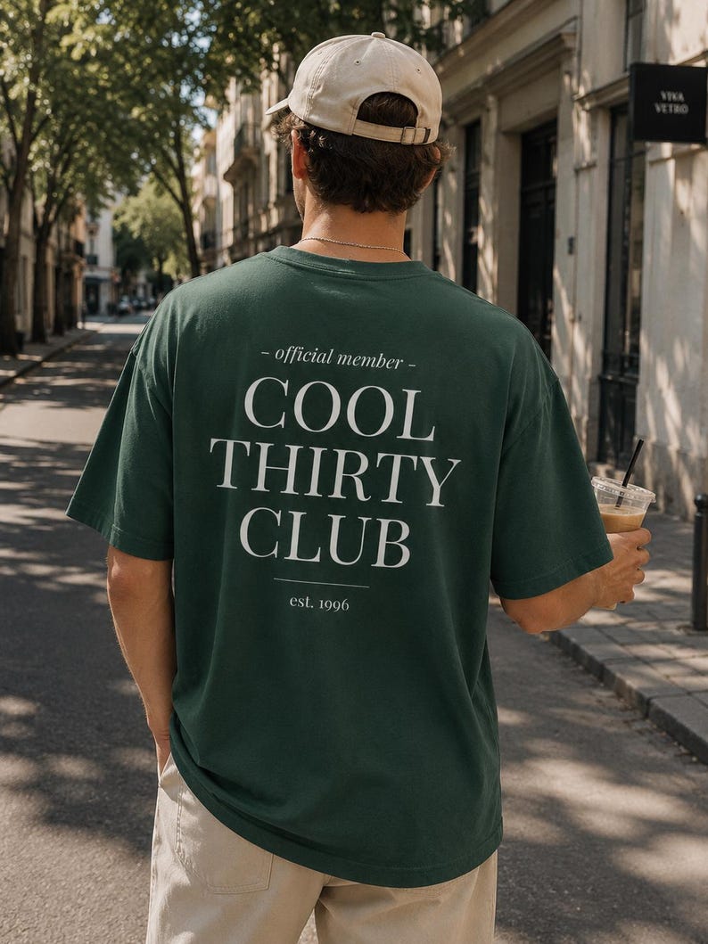 May include: A dark green short-sleeve t-shirt featuring white text on the back that reads - official member - COOL THIRTY CLUB est. 1996. The shirt is paired with a beige baseball cap and light-colored trousers, viewed from the back.