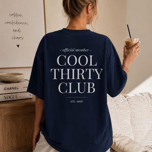 May include: A navy blue short-sleeve t-shirt featuring white text on the back that reads: - official member - COOL THIRTY CLUB est. 1996. The shirt has a crew neckline and a loose, relaxed fit, displaying the back design clearly.