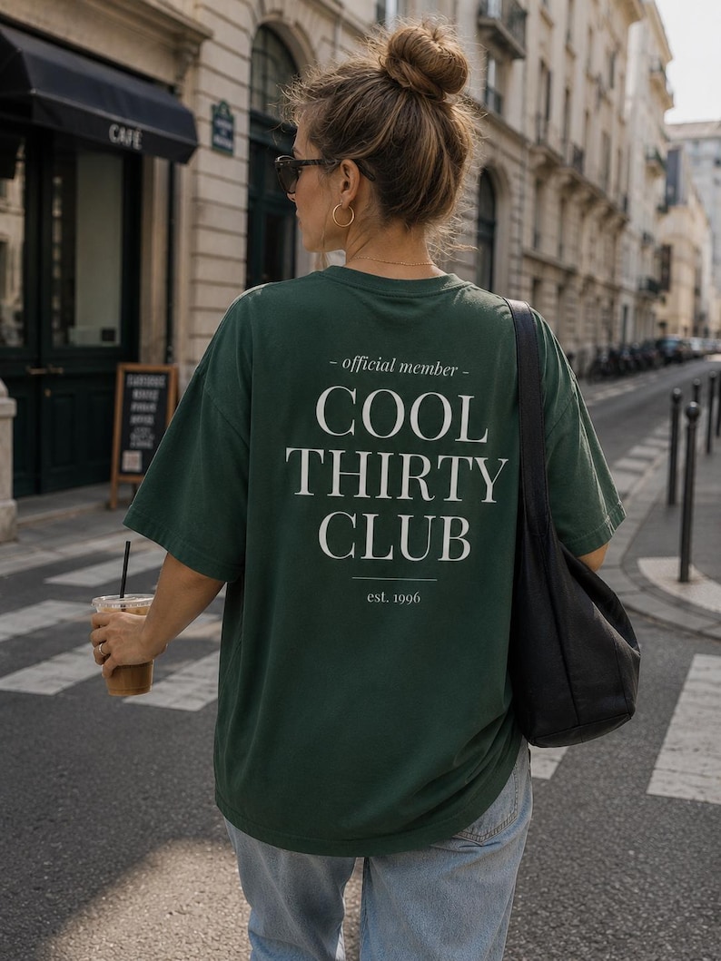 May include: A dark green short-sleeve t-shirt featuring white text on the back that reads - official member - COOL THIRTY CLUB est. 1996. The shirt has a loose, relaxed fit with a crew neckline and is worn with light blue denim jeans.