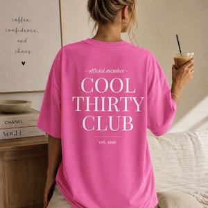 May include: A bright pink short-sleeved t-shirt featuring white text on the back that reads: - official member - COOL THIRTY CLUB est. 1996. The shirt has a loose, relaxed fit with a crew neckline and is worn with light-colored trousers.
