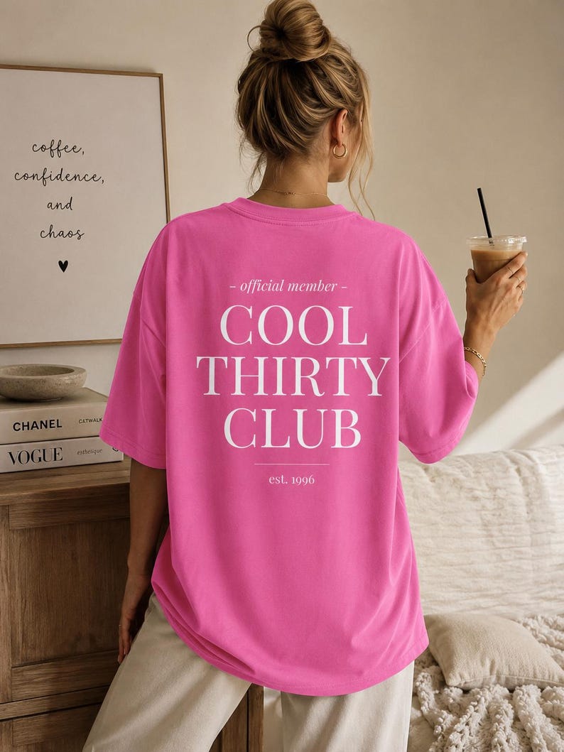May include: A bright pink short-sleeved t-shirt featuring white text on the back that reads: - official member - COOL THIRTY CLUB est. 1996. The shirt has a loose, relaxed fit with a crew neckline and is worn with light-colored trousers.