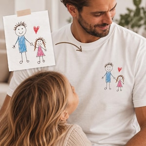 May include: A white t-shirt featuring a small, colourful child's drawing of two stick figures holding hands with a red heart above them. The drawing shows one figure in a blue shirt and one in a pink dress, matching the paper drawing shown above.