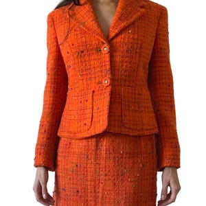 Puede incluir: Traje de falda de tweed naranja. La chaqueta tiene solapas cl&aacute;sicas, botones y dos bolsillos delanteros. La minifalda a juego completa el conjunto. La tela tiene una apariencia texturizada con motas de color.