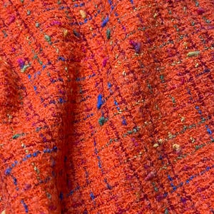Puede incluir: Primer plano de una tela naranja texturizada con motas azules, verdes y rosas. El material tejido tiene un aspecto ligeramente rugoso, lo que sugiere una textura de tweed o boucl&eacute;. Adecuado para ropa o decoraci&oacute;n del hogar.