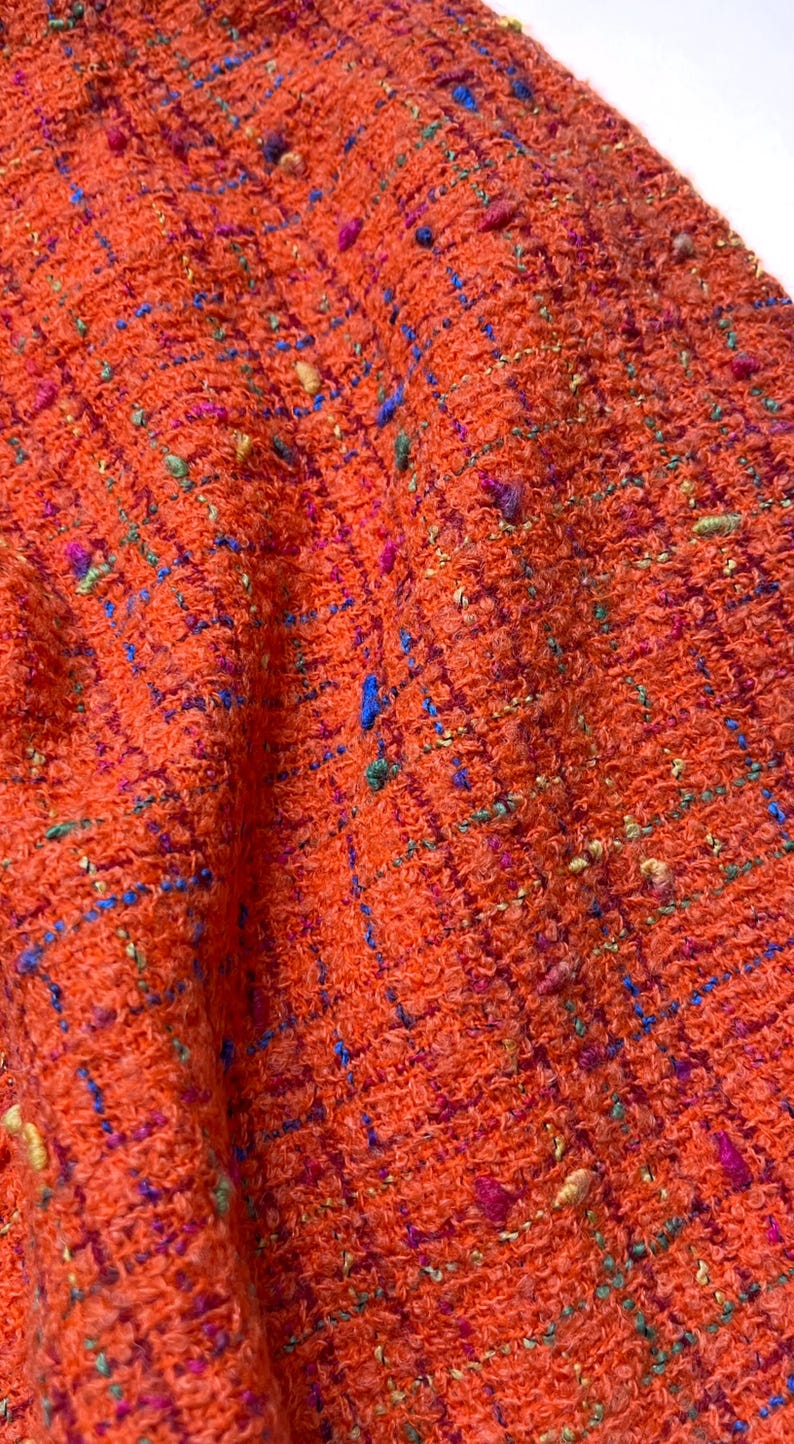 Puede incluir: Primer plano de una tela naranja texturizada con motas azules, verdes y rosas. El material tejido tiene un aspecto ligeramente rugoso, lo que sugiere una textura de tweed o boucl&eacute;. Adecuado para ropa o decoraci&oacute;n del hogar.