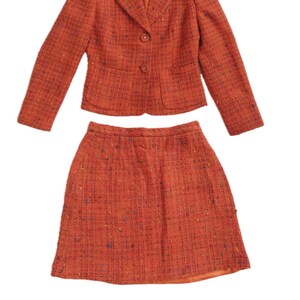 Puede incluir: Traje de dos piezas de tweed naranja. La chaqueta tiene cuello con solapas, dos botones y mangas largas. La falda es de tweed naranja a juego. La etiqueta Versace es visible.