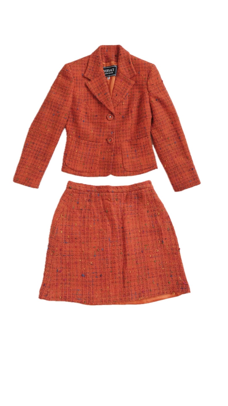 Puede incluir: Traje de dos piezas de tweed naranja. La chaqueta tiene cuello con solapas, dos botones y mangas largas. La falda es de tweed naranja a juego. La etiqueta Versace es visible.