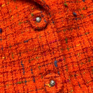 Puede incluir: Primer plano de una chaqueta de tweed naranja con cuello y dos botones decorativos. La tela de tweed tiene motas azules, verdes y rosas. Los botones son redondos con un emblema plateado.