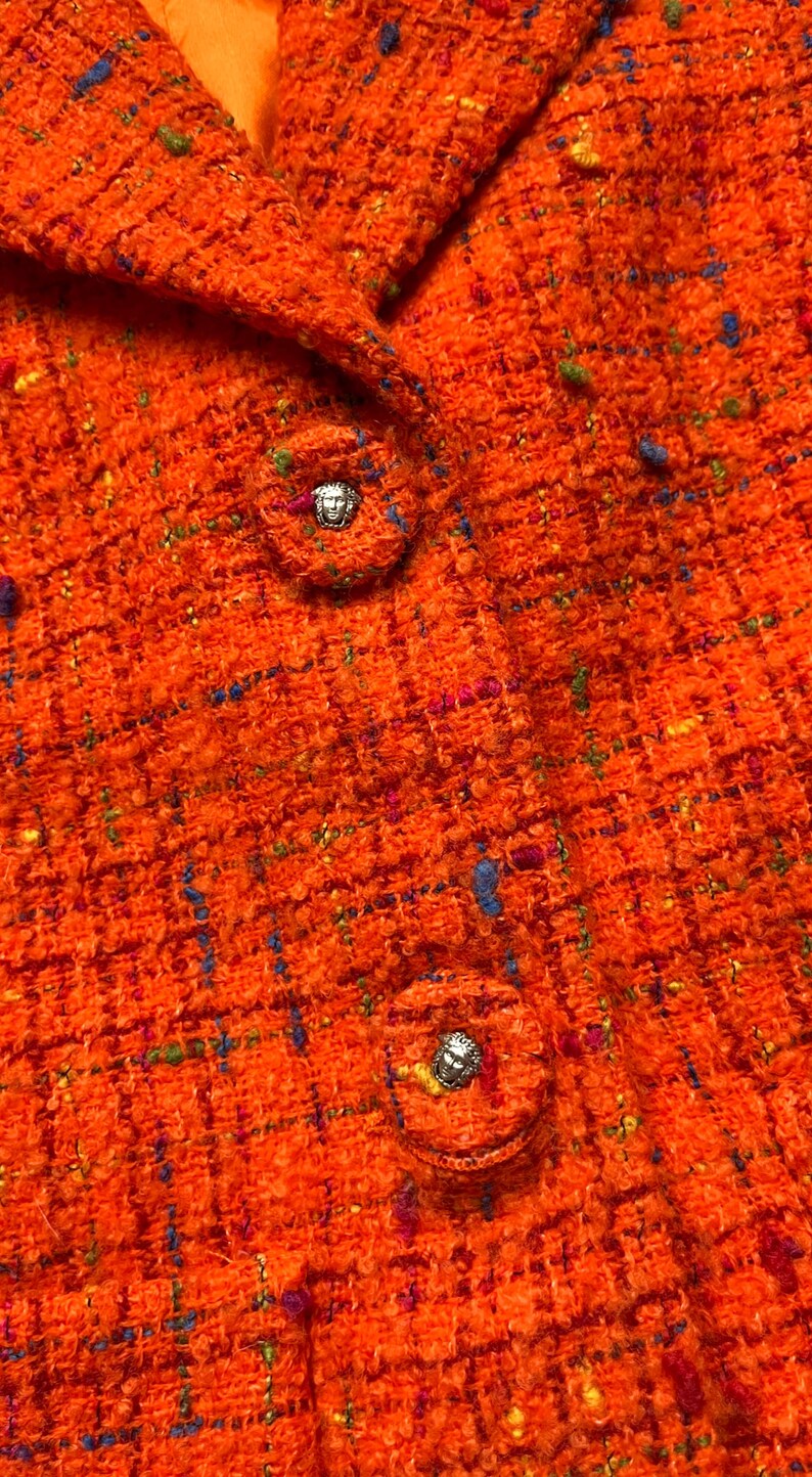Puede incluir: Primer plano de una chaqueta de tweed naranja con cuello y dos botones decorativos. La tela de tweed tiene motas azules, verdes y rosas. Los botones son redondos con un emblema plateado.