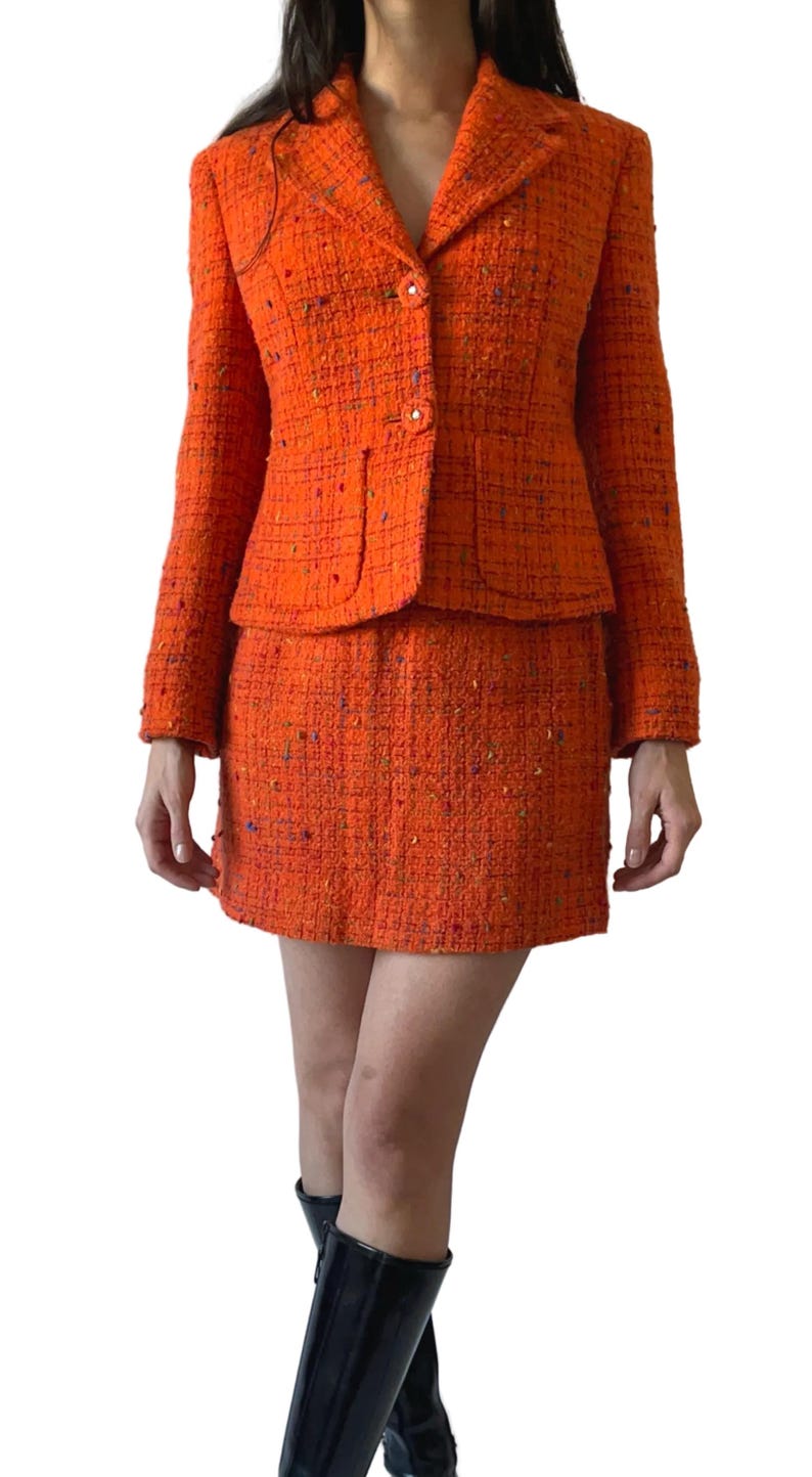Puede incluir: Traje de falda de tweed naranja. La chaqueta tiene solapas cl&aacute;sicas, botones y dos bolsillos delanteros. La minifalda a juego completa el conjunto. La tela tiene una apariencia texturizada con motas de color.