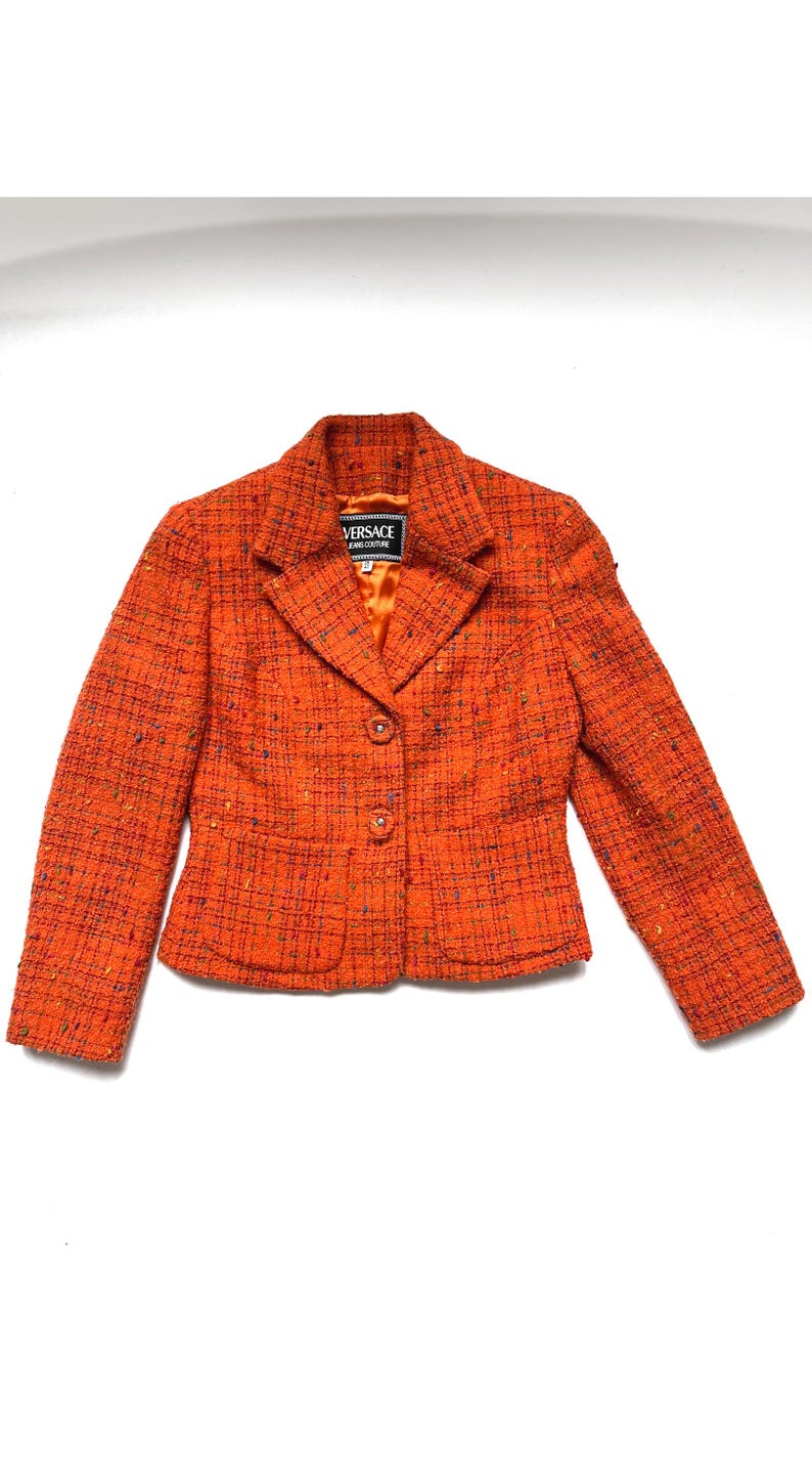 Puede incluir: Blazer de tweed naranja con cierre de dos botones y solapas con muescas. La chaqueta presenta una etiqueta Versace y dos bolsillos delanteros. La tela tiene un aspecto texturizado con motas azules y verdes.