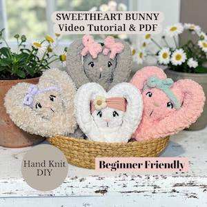 Könnte beinhalten: Eine Sammlung herzförmiger Häschen-Plüschtiere in einem Weidenkorb. Die Häschen sind in verschiedenen Farben, darunter Grau, Beige, Weiß und Rosa, mit Schleifen und floralen Akzenten. Der Text auf dem Bild lautet "Sweetheart Bunny Video Tutorial & PDF", "Hand Knit DIY" und "Beginner Friendly."