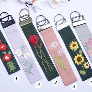 以下が含まれることがあります： 刺繍の花柄が施された6つの布製キーホルダー。左から順に、赤い花のダークグリーン、白い花のライトパープル、赤いバラのダークグリーン、紫の花のピンク、ひまわりのダークグリーン、ピンクの花のライトグレーのキーホルダーが並んでいます。
