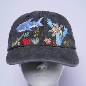 Könnte beinhalten: Eine anthrazitfarbene Baseballkappe mit einer detaillierten Stickerei einer Unterwasserszene. Das Design zeigt einen blauen Hai, eine Meeresschildkröte, rote Korallen, hellblauen Seetang und verschiedene grüne Wasserpflanzen auf der Vorderseite.