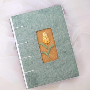 May include: A handmade journal with a sage green linen cover. A rectangular cutout reveals a tan fabric with a yellow and green embroidered flower. The journal is bound with white thread.