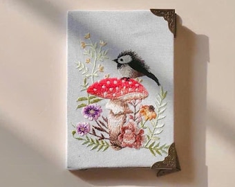 May include: Embroidered artwork featuring a red mushroom with white spots, a small bird, and colorful flowers. The artwork is on a light blue canvas with decorative corner protectors. The embroidery details a whimsical forest scene.