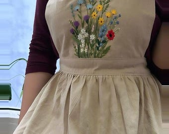 May include: Beige apron with an embroidered floral design in various colors. The apron features a bib top, waist tie, and a pleated skirt. The embroidered flowers include blue, yellow, red, and white blossoms. The apron is made of a light-colored fabric.