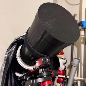 May include: A black telescope with a large cylindrical tube and a red and black base. The telescope is covered with a black protective cover. A small red device is attached to the telescope.