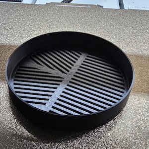 May include: A black circular 3D printed object with a grid pattern on the top. The object is sitting on a textured surface.