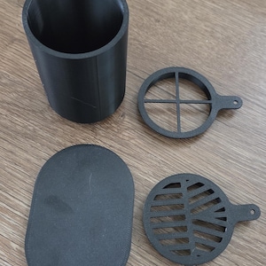 May include: A matte black cylindrical container accompanied by three separate components: a solid oval base plate, a circular insert with a cross-shaped divider, and a circular insert featuring a leaf-like vein pattern, all with small tabs.