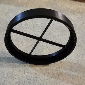 May include: A black, circular object with a cross-shaped interior support structure. The object is made of a solid material and sits on a light-colored, textured surface. The design is simple and geometric.