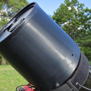 May include: A black telescope with a cylindrical body and a lens at the top. The telescope is mounted on a red base with black straps. The background features green grass, trees, and a glimpse of a white shed under a blue sky.