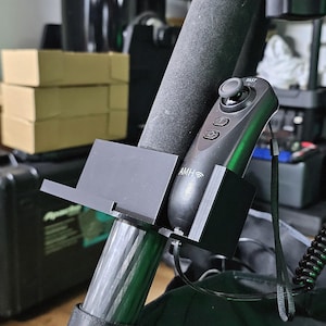 May include: A black remote control with the text "FAST" and the letters "AMH" is mounted on a black bracket attached to a black pole. The remote has several buttons and a joystick. The bracket is attached to a larger black object.