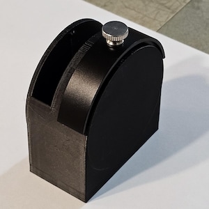 May include: A black 3D printed device with a silver knob on top. The device has a curved shape and a slot on the side.