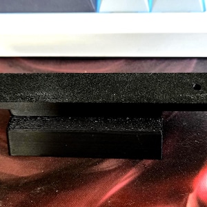 Peut inclure: Un objet noir rectangulaire imprimé en 3D avec une plateforme surélevée et deux petits trous. L'objet est probablement un support ou un support, et il repose sur une surface à motifs rouges et noirs.