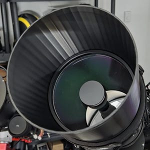 May include: A large black telescope with a wide aperture, featuring a reflective mirror and a secondary mirror. The telescope has a ribbed, cylindrical tube and is equipped with red electronic components. Other telescopes and equipment are visible in the background.