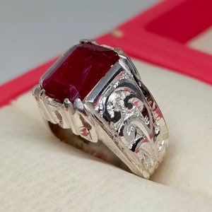 May include: A silver ring with an ornate design and a large, square-shaped, red gemstone.