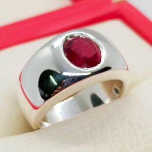 Puede incluir: Un anillo de plata con una gran piedra preciosa de rubí de forma ovalada engastada en el centro. El anillo es grueso y tiene un acabado pulido.