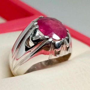 Puede incluir: Un anillo de plata con una gran piedra de rubí rosa de forma ovalada. El anillo tiene un diseño detallado y adornado.