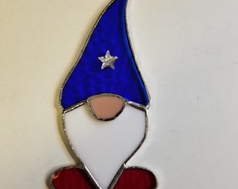 May include: Stained glass gnome with a blue hat featuring a silver star, a white beard, a pink nose, and red shoes. The gnome is outlined in silver and has a loop for hanging. A whimsical decoration.
