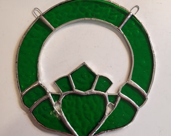 May include: A green stained glass Celtic knot, also known as a Claddagh, in the shape of a heart with two hands holding it. The knot is surrounded by a silver frame.