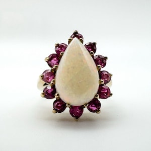 May include: A gold ring featuring a pear-shaped opal center stone surrounded by a halo of small, round, deep pink gemstones. The ring is set against a white background, highlighting the colors and design of the jewelry.