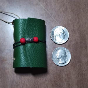 May include: A small, green leather journal with a black cord and red beads wrapped around it. Two quarters are next to the journal. The journal has a textured surface and a string attached.