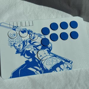 Może przedstawiać: Biały kontroler do gier z niebieską ilustracją wojownika z mieczem. Kontroler ma osiem niebieskich przycisków i d-pad. Projekt tworzy uderzający kontrast na białej powierzchni.