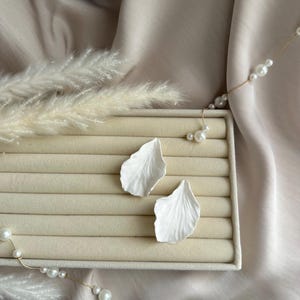 May include: Two white, petal-shaped earrings sit on a beige jewellery display box. The earrings have detailed veining, and are set against a backdrop of soft, draped fabric. A gold chain with pearls is draped around the display.