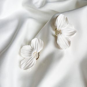 Könnte beinhalten: Ein Paar blumenförmige Ohrringe in Weiß mit texturierten Blütenblättern und einer goldfarbenen, kreisförmigen Mitte, die mit kleinen weißen Perlen verziert ist. Die Ohrringe haben ein zartes, organisches Aussehen mit feinen Rillen.