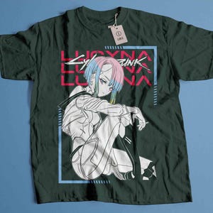 Peut inclure: T-shirt vert fonc&eacute; avec une illustration de style anime. Le motif pr&eacute;sente un personnage aux cheveux bleus et roses, le texte "LUCYNA LUNA" en rose et un cadre rectangulaire bleu. Une &eacute;tiquette indique "LARGE".