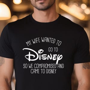 A man wearing a black t-shirt with white text that says "My wife wanted to go to Disney so we compromised and came to Disney".