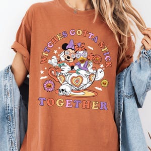 May include: Rust-colored t-shirt with a graphic featuring Minnie Mouse and Daisy Duck in witch costumes, inside a teacup. The text reads "Witches Gotta Stick Together" in a retro font, with stars and floral accents.