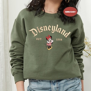 A woman wearing a green sweatshirt with the text "Disneyland EST. 1955" and a Minnie Mouse graphic.