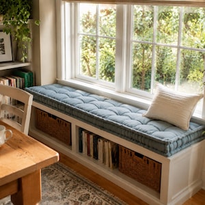 May include: A white window seat bench featuring a long, tufted, dusty blue cushion and a single cream-colored throw pillow. The base of the bench includes open shelving compartments containing books and two woven brown wicker storage baskets.