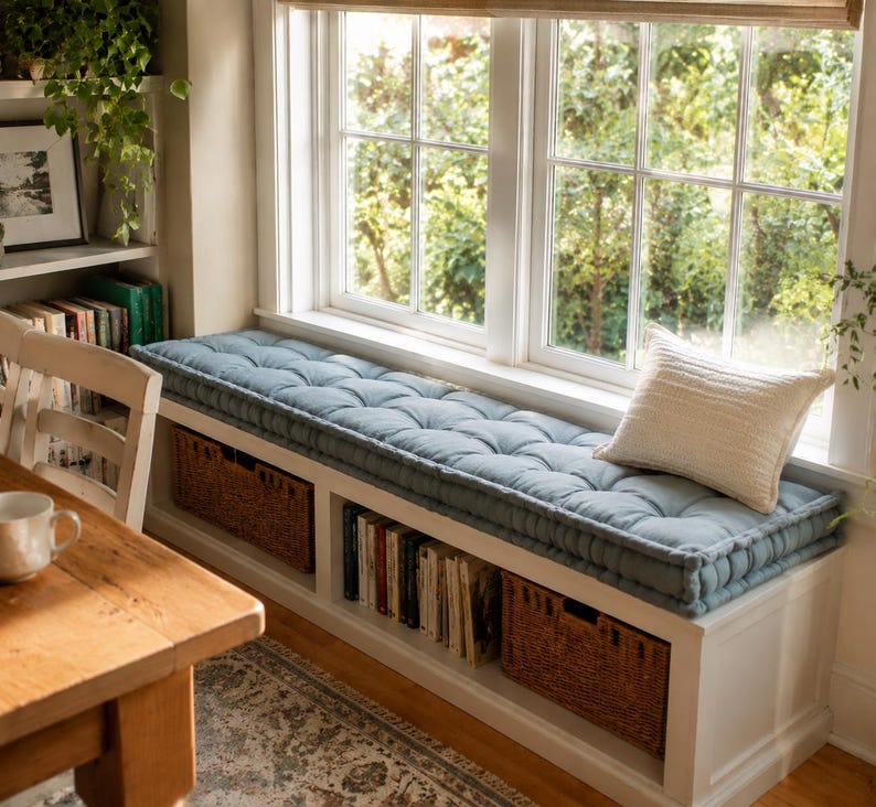 May include: A white window seat bench featuring a long, tufted, dusty blue cushion and a single cream-colored throw pillow. The base of the bench includes open shelving compartments containing books and two woven brown wicker storage baskets.