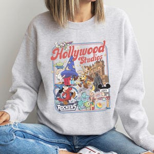 A person wearing a gray sweatshirt with a colorful graphic design featuring various Disney characters and landmarks, such as Mickey Mouse, Minnie Mouse, and the Eiffel Tower.