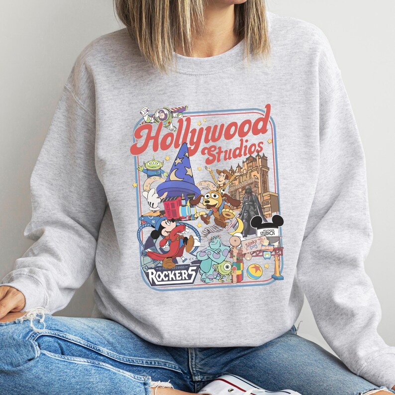 A person wearing a gray sweatshirt with a colorful graphic design featuring various Disney characters and landmarks, such as Mickey Mouse, Minnie Mouse, and the Eiffel Tower.